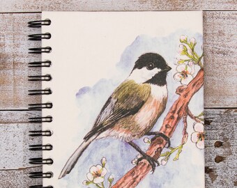 Peut inclure: Un carnet à spirales blanc avec une illustration aquarelle d'une mésange à tête noire perchée sur une branche avec des fleurs blanches. L'oiseau est tourné vers la droite et a un corps brun clair avec une tête noire et un plastron noir.