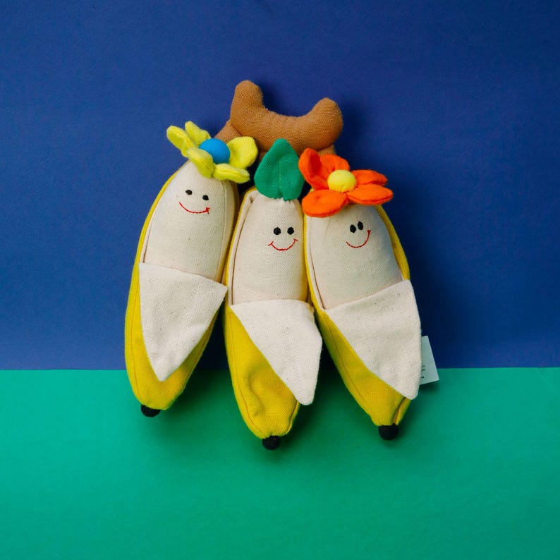 Op de afbeelding: Drie pluche banaan speelgoedjes met lachende gezichten en bloem accenten. De bananen zijn geel met een witte stoffen voering aan de binnenkant. De bloemen zijn oranje, geel en groen.