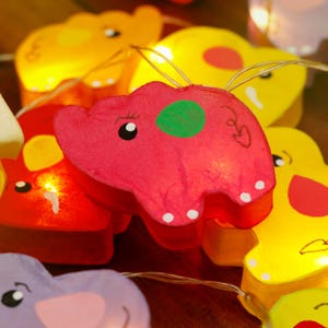 May include: A string of lights with colorful elephant-shaped paper lanterns. The lanterns are red, yellow, purple, and pink with black eyes and white dots. The lights are strung on a clear wire.