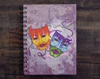 Peut inclure: Un carnet à spirales violet avec une peinture à l'aquarelle de deux masques théâtraux colorés. Les masques sont un masque rouge, jaune et bleu avec une expression souriante et un masque violet, bleu et vert avec une expression triste.