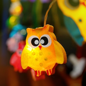 May include: A yellow owl-shaped paper lantern with black eyes and a red beak. The lantern is lit from inside, creating a warm glow.