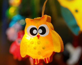 May include: A yellow owl-shaped paper lantern with black eyes and a red beak. The lantern is lit from inside, creating a warm glow.