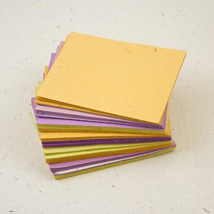 May include: A stack of square-shaped paper sheets in various colors, including yellow, purple, and green. The paper has a textured appearance and is arranged in a slightly fanned-out pattern, showcasing the different hues.