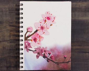 Peut inclure: Un carnet à spirales avec un motif floral rose et blanc sur la couverture. Les fleurs sont des cerisiers en fleurs et l'arrière-plan est un dégradé doux rose et blanc.