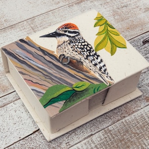 May include: A small, white box with a hand-painted design of a woodpecker perched on a branch with green leaves. The box is made of recycled paper and has a rustic, natural look.