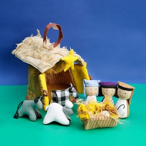 May include: A felt nativity set with a yellow and brown stable, a blue-clad Mary, a purple-clad Joseph, a brown-clad shepherd, a white sheep, a gray donkey, a black and white cow, and a baby Jesus in a manger.