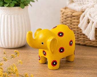 May include: A yellow wooden elephant ornament featuring red and black polka dots. The elephant has a raised trunk and large ears. The ornament is on a wooden surface, with a white vase and a basket in the background. Decorative item.