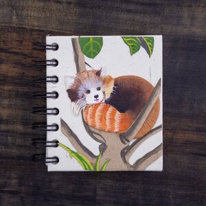 May include: A small spiral bound notebook with a red panda illustration on the cover. The red panda is curled up on a tree branch with green leaves. The notebook has a white background.