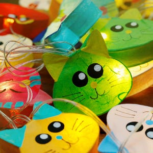 May include: A string of fairy lights with cat-shaped paper lanterns. The lanterns are in various colors, including green, yellow, pink, and blue. Each lantern has a cute cartoon cat face with black eyes and whiskers.
