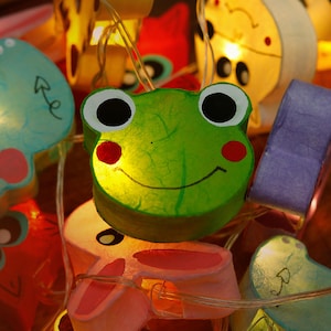 May include: A string of fairy lights with various animal shapes, including a frog, a bunny, a bird, and a cloud. The lights are made of paper and have a warm white glow.
