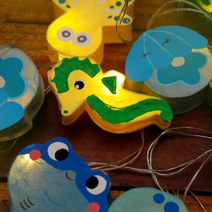May include: A string of fairy lights with wooden animal shapes, including a yellow seahorse, a blue jellyfish, a yellow octopus, a blue flower, and a blue frog. The lights are strung on a clear wire and are lit up.