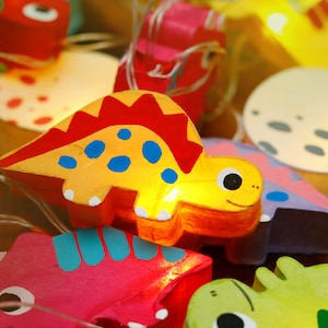 May include: A string of fairy lights with colorful dinosaur-shaped light covers. The dinosaurs are yellow, red, blue, green, and purple. The lights are strung on a clear wire.