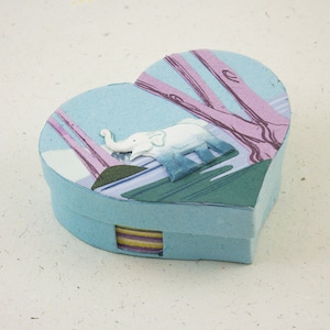 May include: A light blue heart-shaped box with a white elephant design and a colorful striped pattern on the bottom. The elephant is walking through a forest with pink trees.