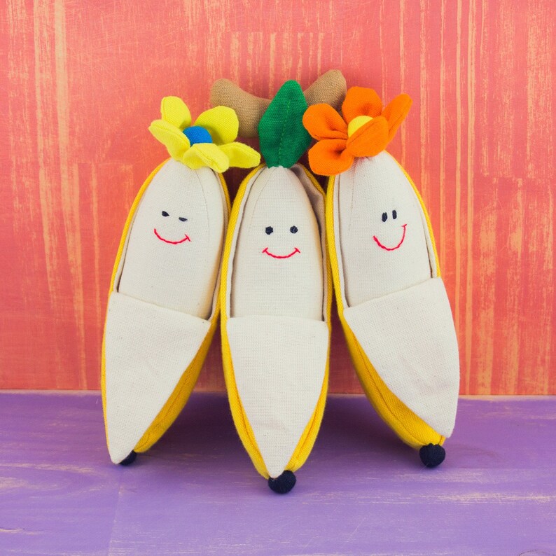 Op de afbeelding: Drie pluche banaan speelgoed met lachende gezichten en bloem accenten. De bananen zijn geel met witte stof en zijn in een rij gerangschikt.