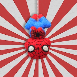 May include: A handmade felt Spider-Man ornament. The ornament features a red head with a black spiderweb design, white eyes, and a blue body. It hangs from a red and white string against a red and white striped background.
