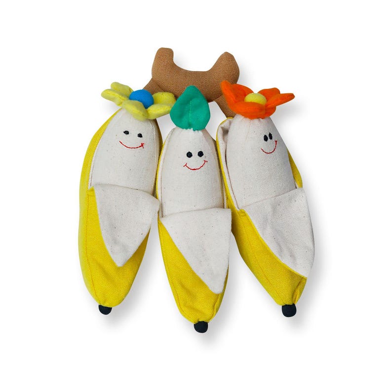 Op de afbeelding: Drie pluche banaan speelgoedjes met lachende gezichten en kleurrijke bloemen accenten. De bananen zijn geel met een witte binnenkant en zijn bevestigd aan een bruine tak.