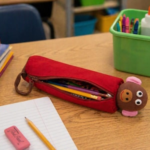 May include: A red pencil case with a brown zipper and a cartoon character attached. The pencil case is open, revealing pencils and pens. An apple, books, a pencil, and an eraser are also on the wooden desk.