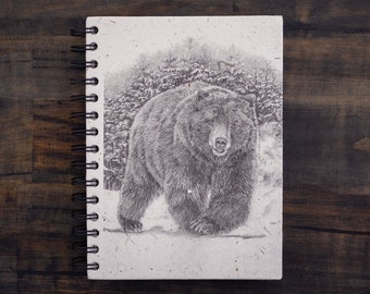 Peut inclure: Un dessin en noir et blanc d'un ours marchant dans une forêt enneigée. Le dessin est sur un carnet à spirales avec une couverture en papier blanc.