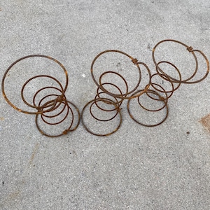 May include: A rusty metal spring structure with multiple circular coils and connecting loops. The coils vary in size, creating an abstract, organic shape. The object is set against a gray concrete surface.