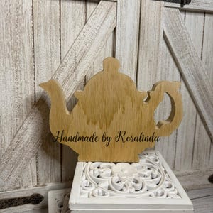 May include: A wooden teapot-shaped cutout with the text "Handmade by Rosalinda" engraved on its surface. The teapot is light brown and sits atop a white, carved box. The background features a white wooden door.