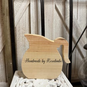 May include: A wooden pitcher-shaped decorative item with a handle. The item is made of light-colored wood, with a darker top section. The text "Handmade by Rosalinda" is engraved on the front. The pitcher is displayed on a white, ornate surface.