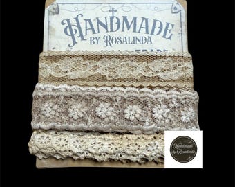 May include: A stack of vintage lace ribbons in various neutral tones, including beige and cream. The ribbons are displayed on a cardboard backing with the text "HANDMADE by Rosalinda". A small, round logo is visible.