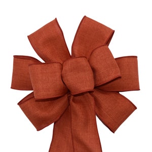 May include: A large, rust-colored bow made from a textured fabric. The bow is tied with a long tail.