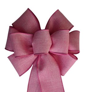 Puede incluir: Un lazo decorativo hecho de tela rosa con una textura tejida visible. El lazo presenta múltiples bucles dispuestos en un patrón simétrico y dos colas largas y rectas que cuelgan hacia abajo. Los bordes están acabados con un borde cosido.