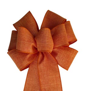 May include: A decorative bow crafted from orange fabric with a visible woven texture. The bow features a central knot with multiple loops radiating outwards and two long, straight tails extending downwards from the centre, all in a solid orange hue.
