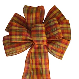 May include: A decorative bow made from fabric with a plaid pattern in shades of orange, yellow, red, and green. The bow features multiple loops and two long, hanging tails that extend downward, showcasing the woven texture of the material.
