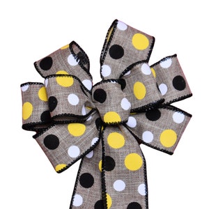 May include: A decorative bow featuring a burlap-textured ribbon with a repeating pattern of yellow, black, and white polka dots. The edges are trimmed with a black stitched border. This bow is suitable for various craft projects.