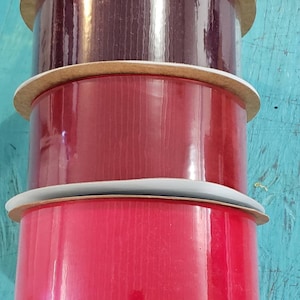 May include: Three spools of ribbon, one red, one burgundy, and one dark red. The spools are stacked on top of each other.