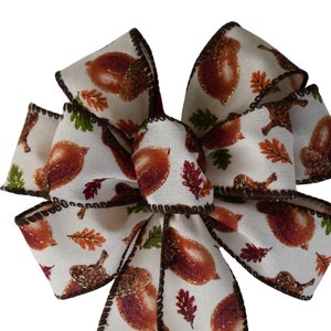 May include: A white fabric bow with a brown trim and a pattern of acorns and leaves. The bow is decorated with gold glitter.