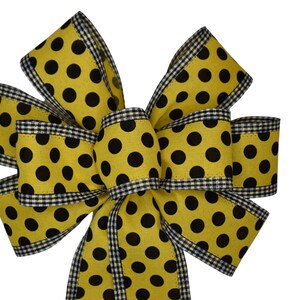 Pode incluir: Um grande laço amarelo com bolinhas pretas e uma borda xadrez preta e branca. O laço é feito de tecido e está amarrado em forma de laço clássico.