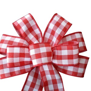 May include: A decorative bow featuring a red and white gingham pattern. The bow is made of fabric and has multiple loops and tails. The edges of the bow are finished with a red trim. The bow is suitable for various craft projects.