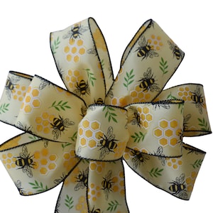 May include: A yellow and black bee-themed bow with a white background. The bow is made of ribbon with a black trim and features a repeating pattern of bees, honeycombs, and green leaves.