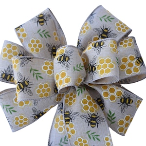 Op de afbeelding: Een beige jute strik met een honingraat- en bijenpatroon. De strik is gebonden met een lus en heeft een rustieke, boerderijstijl.