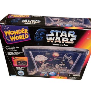 Puede incluir: Una caja de juego de Star Wars Wonder World. La caja es negra con el logotipo de Star Wars y la imagen de Darth Vader. El juego incluye un recipiente de plástico transparente con escenas de batalla de Star Wars. El texto en la caja dice: "¡Convierte el agua en un universo de creatividad!"