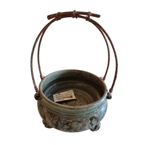 May include: A handcrafted ceramic pot with a copper wire handle. The pot is a muted green with brown accents and rests on three small feet. A small tag is inside the pot.
