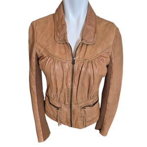 May include: A tan leather jacket with a zip-up front and two front pockets. The jacket has a collared neckline and long sleeves. The jacket is displayed on a white mannequin.