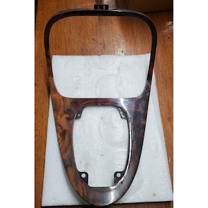 Könnte beinhalten: Ein braunes Holzfurnier-Autoinnenraum-Zierteil mit einer rechteckigen Öffnung in der Mitte. Das Zierteil hat vier schwarze Schraubenlöcher um die Öffnung herum.