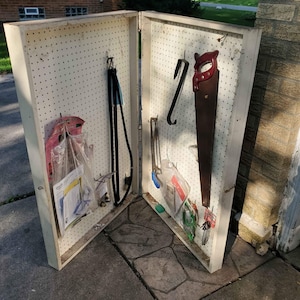 May include: A white pegboard with various tools and supplies hanging on it. The tools include a saw, a wrench, a measuring tape, and a few other items. The pegboard is mounted on a wall and is in a garage or workshop.