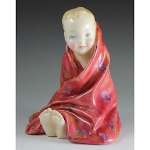 Könnte beinhalten: Eine Porzellanfigur eines Babys, das mit einer roten und orangefarbenen, gemusterten Decke umwickelt sitzt. Das Baby hat einen hellen Teint und blaue Augen.