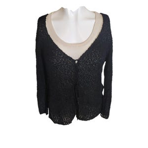 May include: A black, open-weave cardigan with long sleeves, layered over a beige scoop-neck top. The cardigan has a single button closure at the bust. The clothing is displayed on a white mannequin.