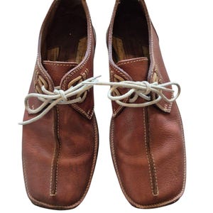 Pode incluir: Um par de sapatos de couro castanho com costuras brancas e atacadores de cor clara. Os sapatos têm bico arredondado e salto baixo. O couro parece desgastado, sugerindo um estilo vintage. Os atacadores estão amarrados.
