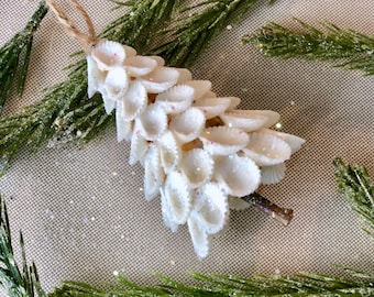 May include: A handmade Christmas ornament crafted from white seashells, arranged in a cascading pattern. The ornament is adorned with iridescent glitter and has a twine loop for hanging. Green pine branches surround the ornament.
