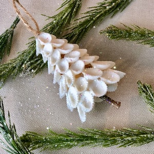 May include: A handmade Christmas ornament crafted from white seashells, arranged in a cascading pattern. The ornament is adorned with iridescent glitter and has a twine loop for hanging. Green pine branches surround the ornament.