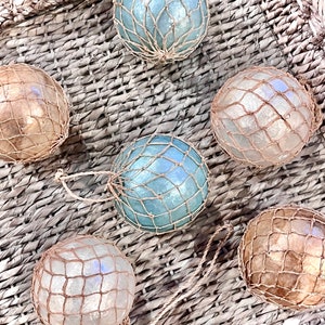 May include: A collection of six spherical glass floats, each encased in a decorative tan jute netting. The spheres feature iridescent finishes in shades of light blue, soft white, and warm tan, arranged in a cluster to display their varied colors.