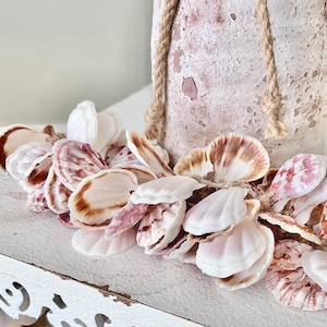 May include: A decorative seashell garland with a variety of colors, including white, pink, and brown, is draped across a white, distressed tray. A textured, light pink vase with a rope handle is in the background.