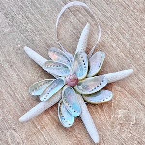 May include: A decorative ornament featuring a flower-like design made from iridescent abalone shells and white starfish. A small, pinkish-purple shell is at the center. A white ribbon is attached for hanging. The ornament is on a wooden surface.